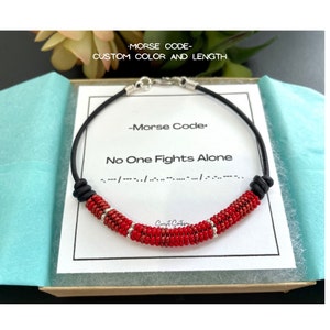 May include: Red beaded bracelet with black cord and silver accents. The bracelet spells out "No One Fights Alone" in Morse code.