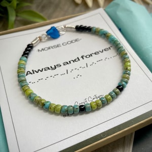 Peut inclure: Un bracelet en perles turquoise, vert et noir avec un charme argenté et un petit cœur bleu. Le bracelet est dans une boîte avec une carte blanche qui dit "Code Morse" et "Toujours et à jamais" en code Morse.