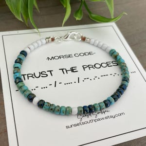 May include: A beaded bracelet with white, turquoise, and navy blue beads. The bracelet is on a white card with the words "TRUST THE PROCESS" in Morse code. The bracelet has a silver clasp.