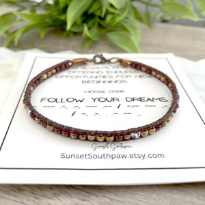 Peut inclure: Un bracelet en perles brunes et rouges avec des accents argentés. Le bracelet est sur une carte blanche avec le texte "FOLLOW YOUR DREAMS" en lettres noires et le code morse de la phrase en dessous. La carte comprend également le texte "Sunset Southpaw.etsy.com".