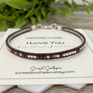 Peut inclure: Un bracelet en cuir marron avec des perles blanches et bordeaux qui épellent "Je t'aime" en code Morse. Le bracelet est sur une carte blanche avec le texte "Code Morse - Je t'aime" et l'adresse du site web "sunsetsouthpaw.etsy.com".