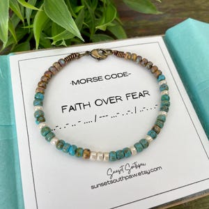 May include: A turquoise and brown beaded bracelet with a silver clasp. The bracelet has a card attached to it that says "Morse Code - Faith Over Fear" and the Morse code for the phrase.