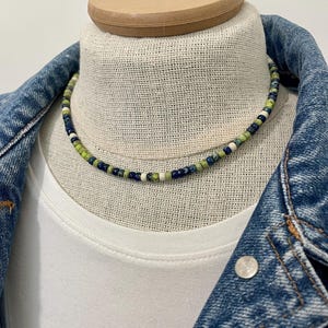 May include: A beaded necklace with a mix of green, blue, white, and dark blue beads. The necklace is displayed on a mannequin head, layered over a white t-shirt and a denim jacket.