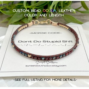 May include: A brown leather bracelet with gold beads and red, blue, and gold beads. The bracelet has the text "Dont Do Stupid Shit" and "MORSE CODE" on a white card. The text "--/--/..../..." is also on the card. The bracelet is from the shop "Sunset Southpaw" on Etsy.