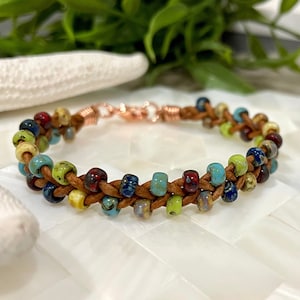May include: A braided leather bracelet with colorful glass beads. The beads are in shades of blue, green, yellow, and red. The bracelet has a copper clasp.