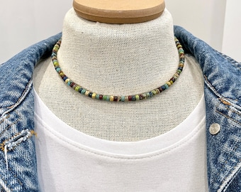 May include: A multi-colored beaded necklace with a variety of blue, green, and brown beads. The necklace is on a white mannequin wearing a white t-shirt and a denim jacket.