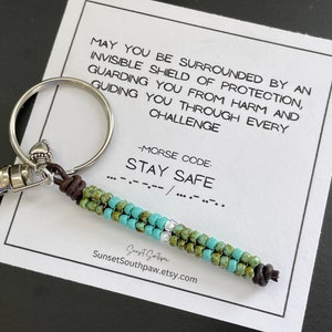 May include: A keychain with a beaded charm in shades of green, turquoise, and silver. The charm is attached to a silver clasp and a brown leather cord. A card with the text "STAY SAFE" in Morse code is in the background.
