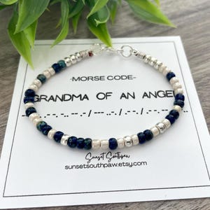 May include: A beaded bracelet with white and blue beads and silver accents. The bracelet is on a white card with the text "MORSE CODE - GRANDMA OF AN ANGEL" and a morse code translation.