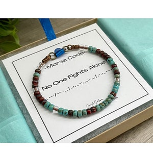 May include: A beaded bracelet featuring alternating turquoise, deep red, and clear glass beads. The bracelet includes copper-colored wire accents and a blue heart-shaped charm. It rests on a white card printed with the text No One Fights Alone.