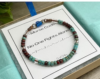May include: A beaded bracelet featuring alternating turquoise, deep red, and clear glass beads. The bracelet includes copper-colored wire accents and a blue heart-shaped charm. It rests on a white card printed with the text No One Fights Alone.