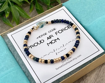 Puede incluir: Una pulsera de cuentas que alterna cuentas azul oscuro y dorado metálico, con un pequeño dije azul claro. La pulsera descansa sobre una tarjeta blanca impresa con el texto MORSE CODE PROUD AIR FORCE MOM y los puntos y rayas del código Morse.