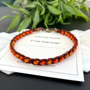 May include: A brown leather bracelet with orange and brown beads. The bracelet is on a white card with the text "Morse Code for Cancer" and a website address.
