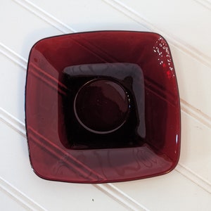 May include: A square, dark red glass saucer with a slightly raised center. The saucer has a subtle, striped pattern.