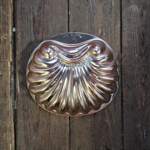 May include: A vintage, silver-colored metal seashell-shaped mold. The mold has a detailed, ridged design and a circular shape. The mold is likely used for baking or creating decorative food items. The mold is sitting on a wooden surface.