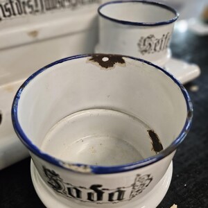 May include: White enamelware soap dish and cup set with blue trim. The soap dish has the word "Soda" in black lettering. The cup has the word "Seife" in black lettering. The set is vintage and shows signs of wear.