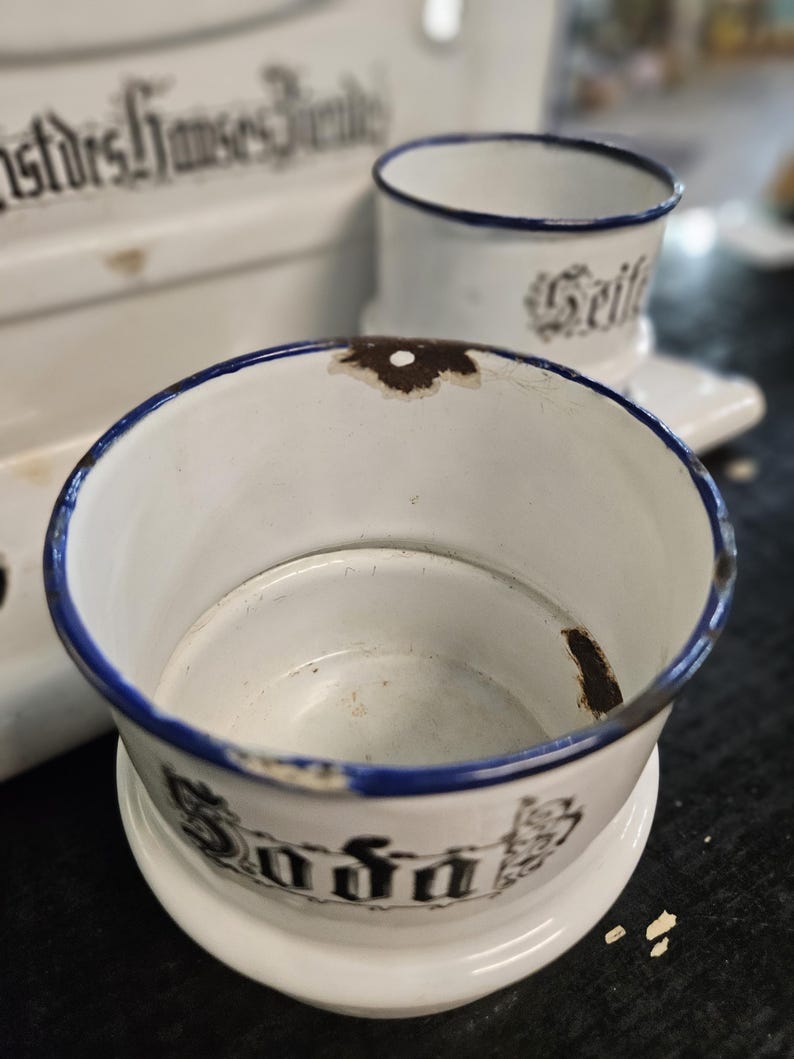 May include: White enamelware soap dish and cup set with blue trim. The soap dish has the word "Soda" in black lettering. The cup has the word "Seife" in black lettering. The set is vintage and shows signs of wear.