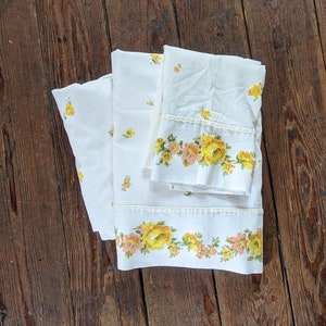 May include: A set of white bed linens with a floral pattern featuring yellow and pink roses. The linens have a lace trim.