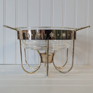 May include: A gold-toned metal chafing dish with a clear glass bowl and a star-shaped design around the rim. The dish has a round base with four legs.