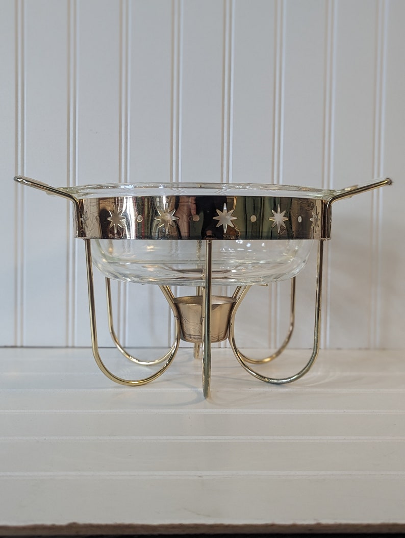 May include: A gold-toned metal chafing dish with a clear glass bowl and a star-shaped design around the rim. The dish has a round base with four legs.