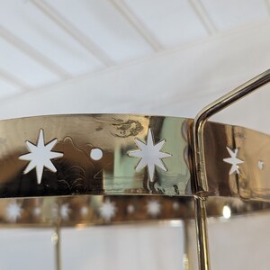 May include: Gold metal stand with a round top and three legs. The top of the stand has a decorative design of white stars.