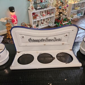 May include: A white enamel kitchen utensil holder with a blue trim and three circular openings. The text "Ordnung ist des Hauses Zierde" is printed on the top. The holder is on a dark surface.