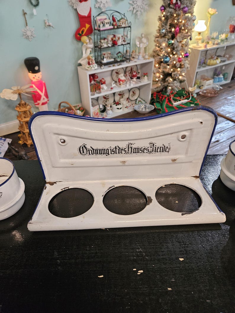May include: A white enamel kitchen utensil holder with a blue trim and three circular openings. The text "Ordnung ist des Hauses Zierde" is printed on the top. The holder is on a dark surface.