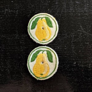 May include: Two embroidered fabric patches with a yellow pear design on a white background. The pear has a green stem and leaves. The patches are surrounded by a green circle.