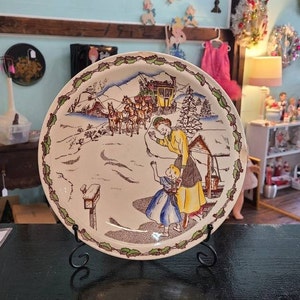 Peut inclure: Une assiette en céramique vintage avec une scène d'hiver représentant une femme et un enfant dans un paysage enneigé. L'assiette a un fond blanc avec des accents bruns, verts et rouges. La scène représente une femme en robe jaune et un enfant en robe bleue marchant dans la neige. Il y a des chevaux et une calèche en arrière-plan.