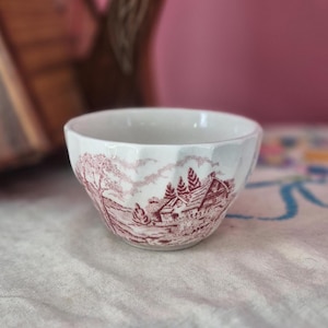 May include: A white ceramic bowl with a faceted exterior and a maroon-colored transferware print depicting a rural landscape with a house, trees, and rolling hills. The bowl features a smooth interior and a scalloped rim design around the top edge.