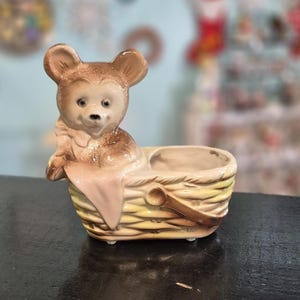 以下が含まれることがあります： 編みかごプランターに入ったセラミック製のクマの置物。クマは明るい茶色で、可愛らしい表情をしています。かごは黄色とタンで、茶色の持ち手が付いています。ピンク色の布がバスケットの縁にかけられています。