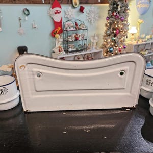 May include: A white enamelware sink with a curved design, featuring a black trim. The sink is surrounded by small white and blue containers with text. The background includes holiday decorations and a Christmas tree.