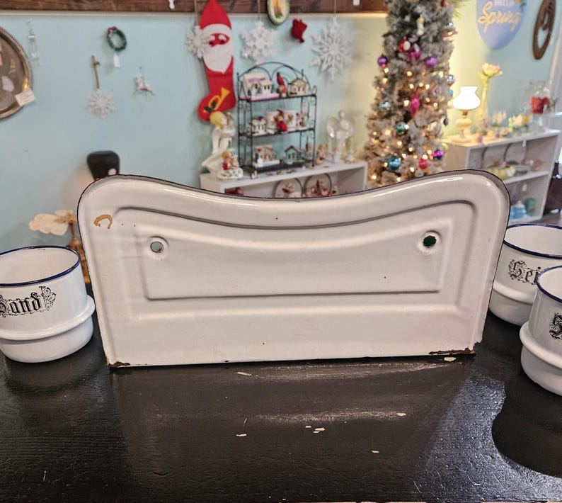 May include: A white enamelware sink with a curved design, featuring a black trim. The sink is surrounded by small white and blue containers with text. The background includes holiday decorations and a Christmas tree.