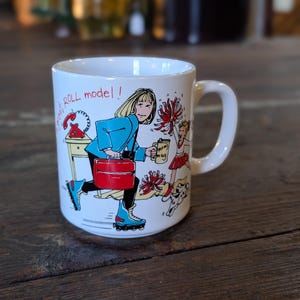 May include: White ceramic mug with a cartoon illustration of a woman roller skating, carrying a briefcase and a coffee mug. The mug has the text "great ROLL model !!" and a drawing of a phone, a cheerleader, and a dog.