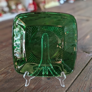 Può includere: Un piatto da portata quadrato in vetro verde smeraldo con una sezione centrale rialzata e un design radiale. Il piatto è esposto su un supporto in plastica trasparente, che mette in risalto la sua estetica vintage. Viene probabilmente utilizzato per servire antipasti o snack.