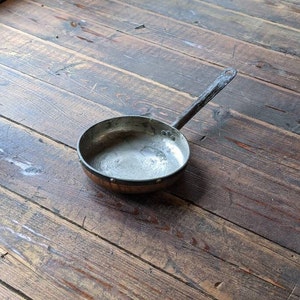 May include: A vintage copper frying pan with a black metal handle. The pan has a worn, rustic look and is sitting on a wooden surface.