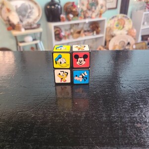 May include: A black cube with six different images of Disney characters. The characters are Mickey Mouse, Donald Duck, Goofy, Pluto, and two images of Mickey Mouse.