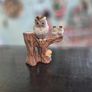 May include: A ceramic owl figurine perched on a tree stump, featuring a large owl and two smaller owls. The figurine is painted in shades of brown, gray, and white, with a small mushroom detail at the base. The owl's eyes are detailed.