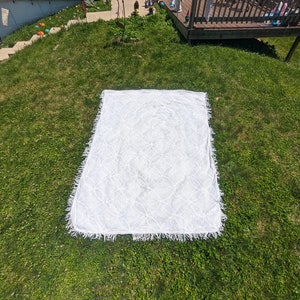 May include: A white crocheted throw blanket with a fringed border, laid out on a green grassy lawn.