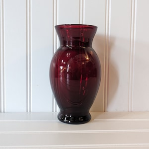 Pode incluir: Um vaso de vidro vermelho rubi com a parte superior alargada e um corpo arredondado. O vaso tem ranhuras verticais e assenta numa base pequena e escura. O fundo é uma parede branca com painéis verticais.