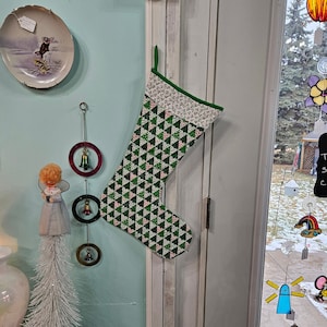 Puede incluir: Una media de Navidad verde y blanca con un patrón geométrico de triángulos. La media está colgada en una puerta blanca.