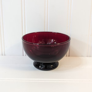 Puede incluir: Un cuenco de vidrio rojo oscuro con un cuerpo redondeado y una base pequeña y circular. El cuenco tiene una superficie lisa y brillante y está colocado sobre un fondo blanco. Probablemente se utiliza para servir postres o porciones pequeñas.