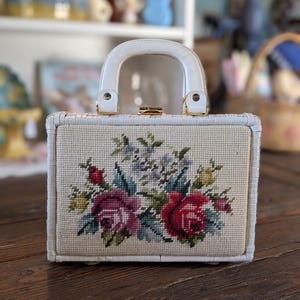 Puede incluir: Bolso de mano blanco vintage con marco tejido y asa blanca. La parte delantera presenta un diseño floral de punto de cruz con rosas rojas y moradas, hojas verdes y flores azules. Tiene un cierre dorado.