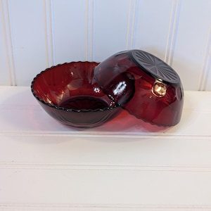 May include: Two ruby red glass bowls. One bowl is upright, and the other is resting on its side. The bowls have a scalloped edge and a textured design. The bowls are on a white surface.