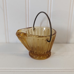 May include: A small amber glass bucket with a wire handle. The bucket has a ribbed design and a rounded bottom.