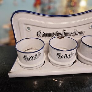May include: A white enamel kitchen organizer with three cylindrical containers labeled "Sand" and "Soda" in black script. The organizer has a blue trim and the text "Ordnung ist des Hauses Zierde" in black.