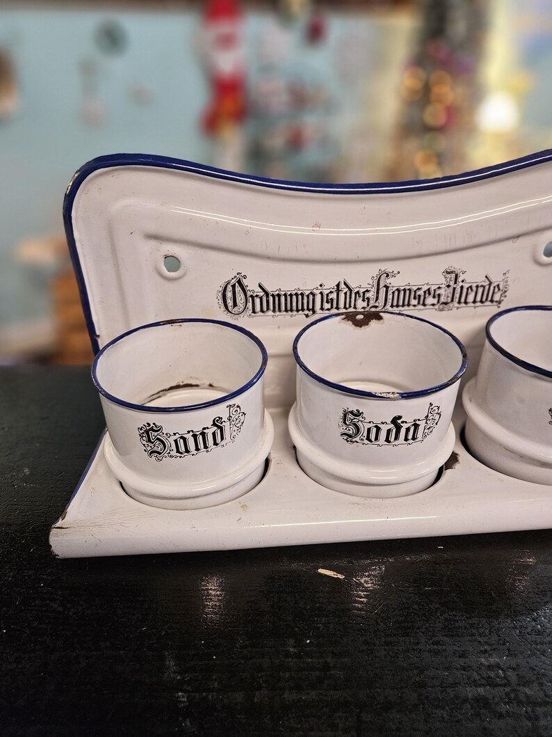 May include: A white enamel kitchen organizer with three cylindrical containers labeled "Sand" and "Soda" in black script. The organizer has a blue trim and the text "Ordnung ist des Hauses Zierde" in black.