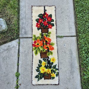 May include: A rectangular latch hook wall hanging featuring a cream-colored background with three vertically stacked brown hanging planters. The pots contain vibrant red, orange, and yellow flowers with green leaves, all rendered in a textured yarn.