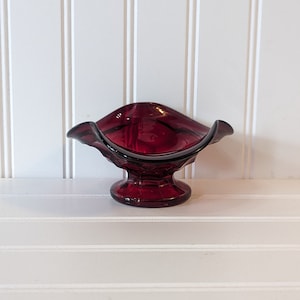 Puede incluir: Un pequeño plato de vidrio rojo rubí con un borde festoneado y una base de pedestal. El plato es perfecto para contener objetos pequeños como joyas o dulces.