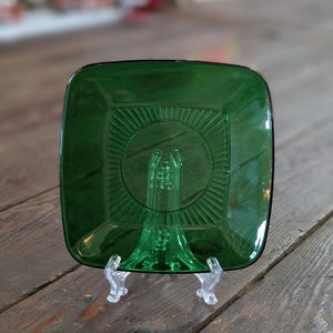 Può includere: Un piatto quadrato di vetro verde smeraldo con un design circolare centrale e linee radiali. Il piatto è esposto su un supporto di plastica trasparente, che ne mette in risalto i dettagli. Il piatto è probabilmente vintage o antico.
