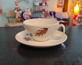 May include: A white teacup and saucer with a brown leaf design. The cup has a rounded shape and a handle. The saucer is round and has a slightly raised edge.